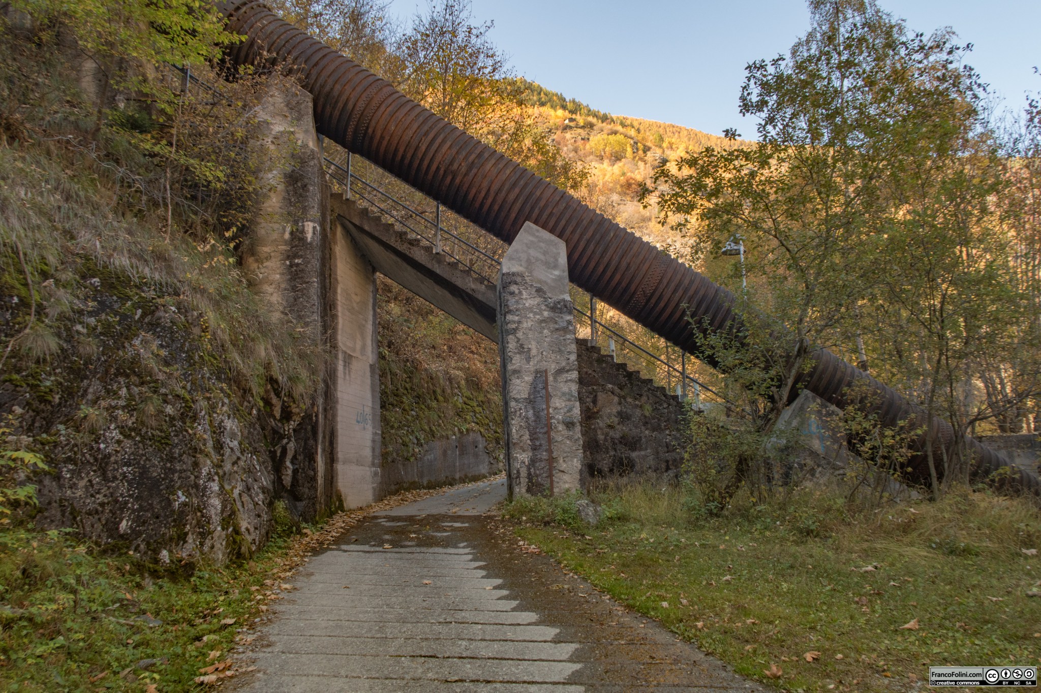 La stradina che rapidamente sale da Vedello verso l'abitato di Ambria in questo tratto passa sotto le condotte forzate che scendono dalla valle e lago di Zappello. 