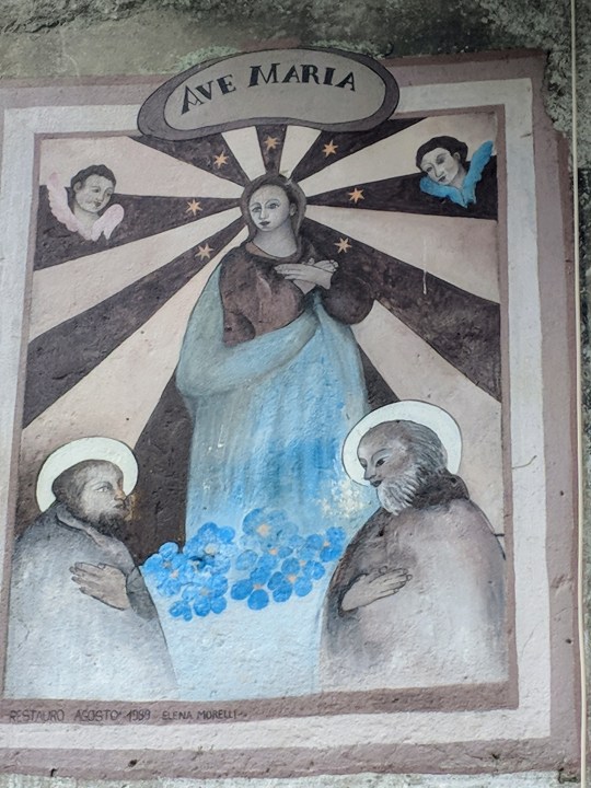 Affresco votivo. Due santi in adorazione della vergine Maria. Iscrizione: “Ave Maria, Restauro Agosto 1969, Elena Morelli”. Il restauro sembra aver danneggiato l’affresco originale e aver reso irriconoscibili i due santi.